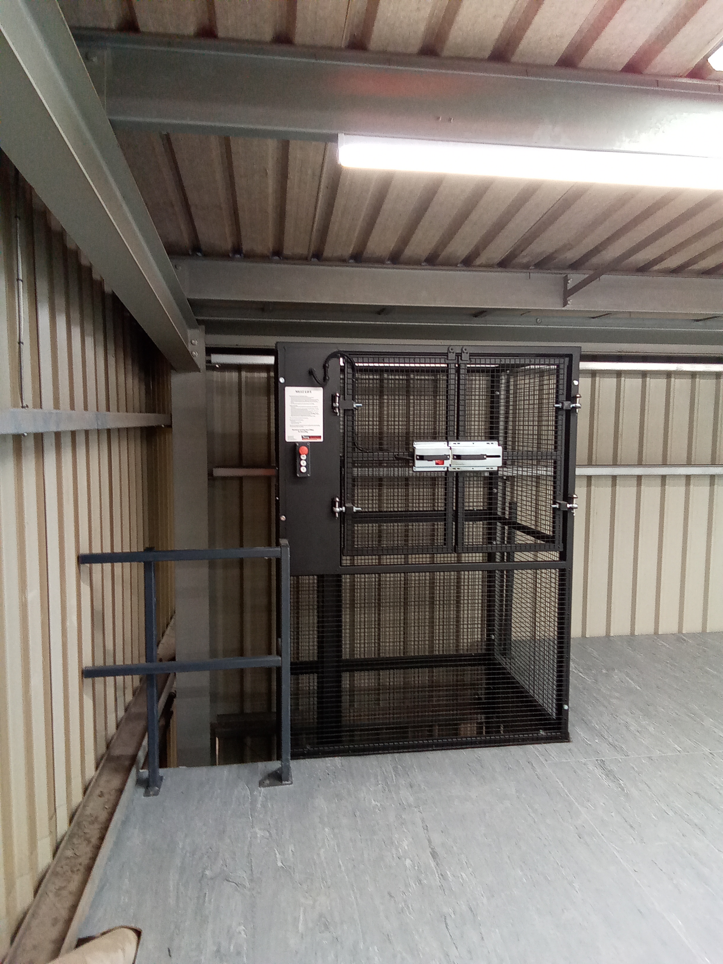 A black metal cage lift with a control panel and safety instructions is installed on an upper floor next to a staircase in an industrial warehouse with metal walls and ceiling.