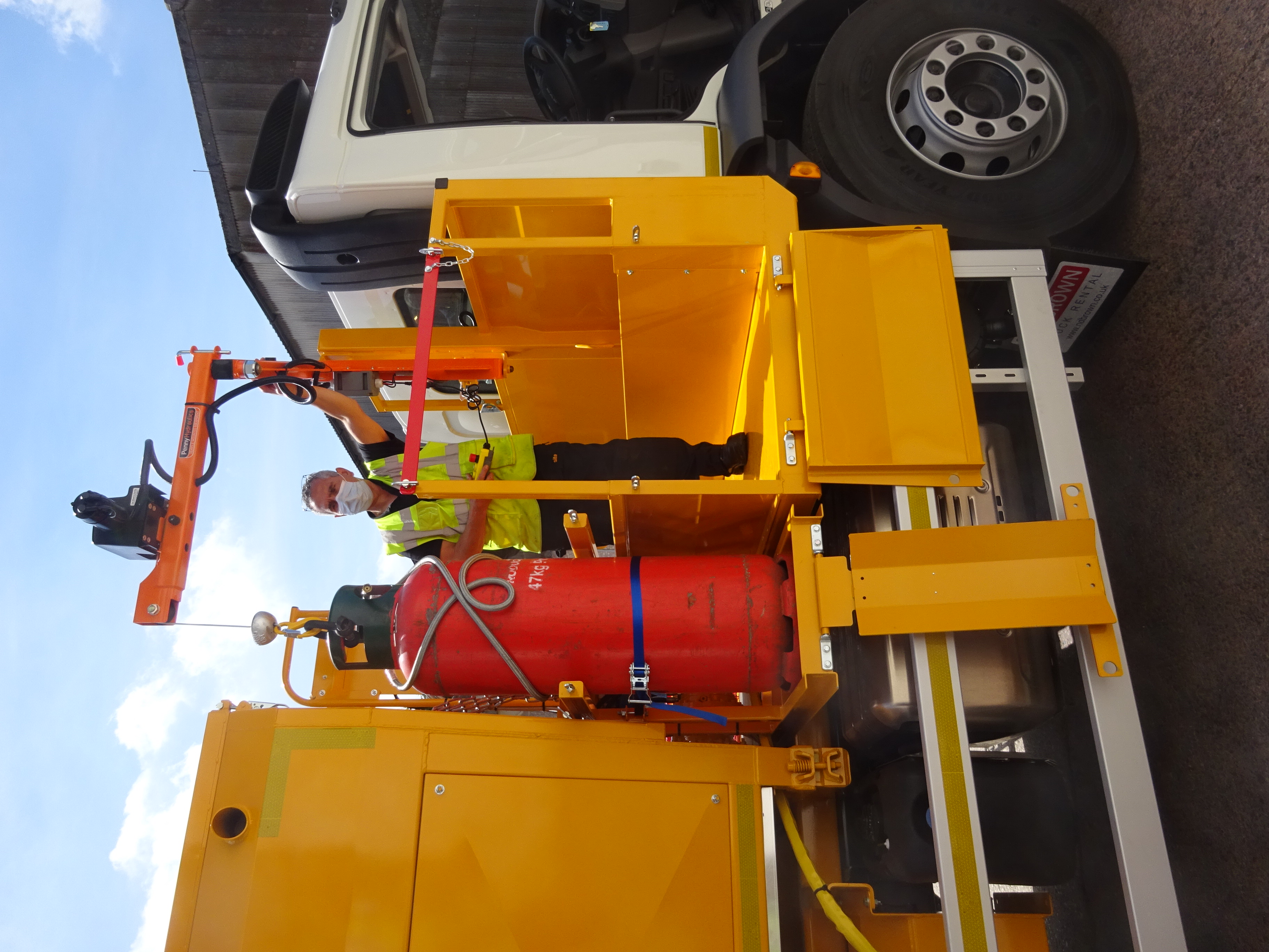 A worker in a high-visibility jacket and face mask uses a lifting device to move a large red gas cylinder onto a platform attached to a yellow utility lorry.