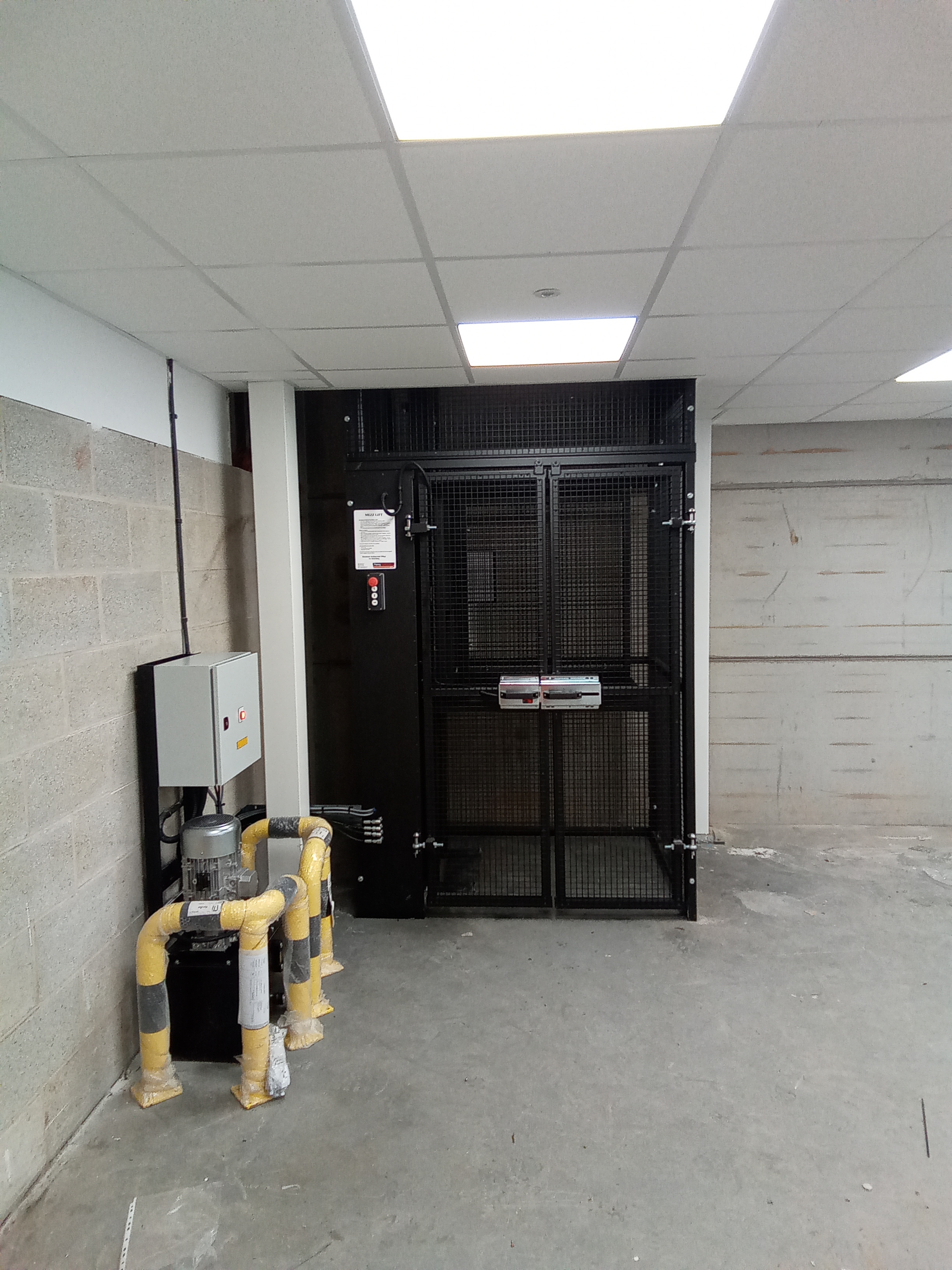 A goods lift with a black metal gate is installed in a basement or industrial area with concrete walls and exposed utility pipes wrapped in yellow and black tape. The floor is unfinished and the ceiling has fluorescent lighting.