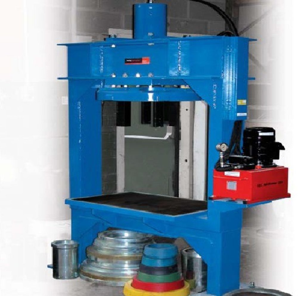 A blue hydraulic press machine with a red control box stands indoors. Several metal rings and colourful cone-shaped objects are placed beside and beneath the press.
