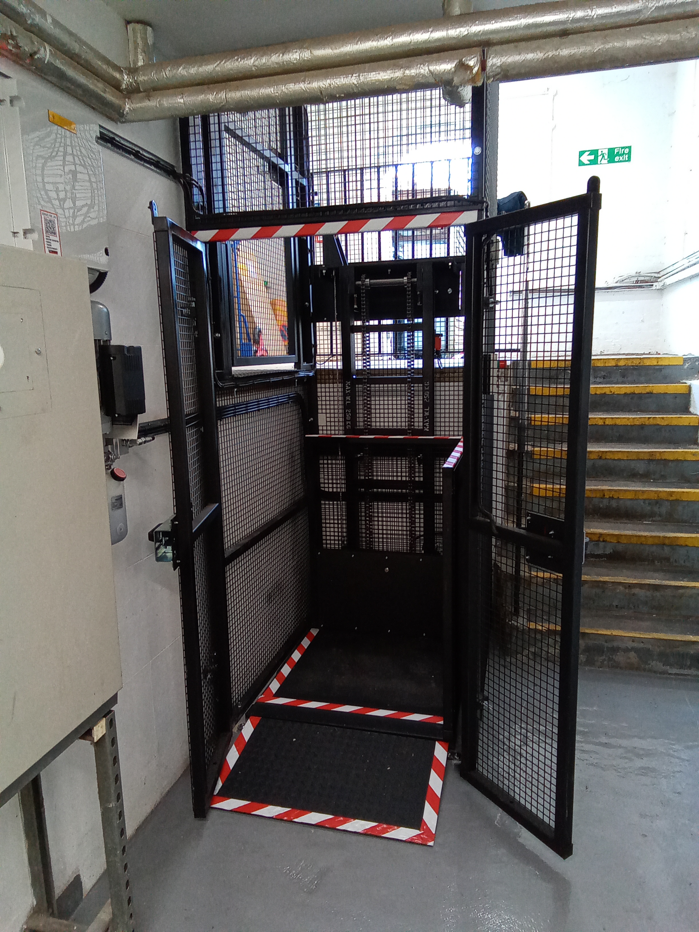 A black metal goods lift with mesh sides and open doors is positioned next to a flight of yellow-edged concrete stairs in an industrial building. Red and white safety tape marks the lift's floor boundary.