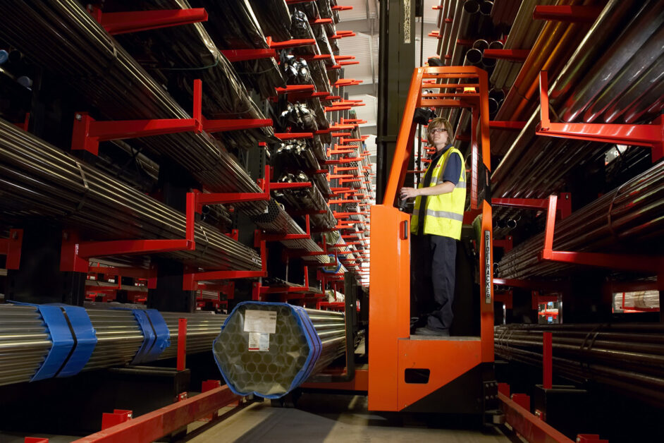 A worker in a yellow safety vest operates an orange lift between racks of long metal pipes stored in a warehouse. The shelves are filled with stacked pipes wrapped in blue bands.