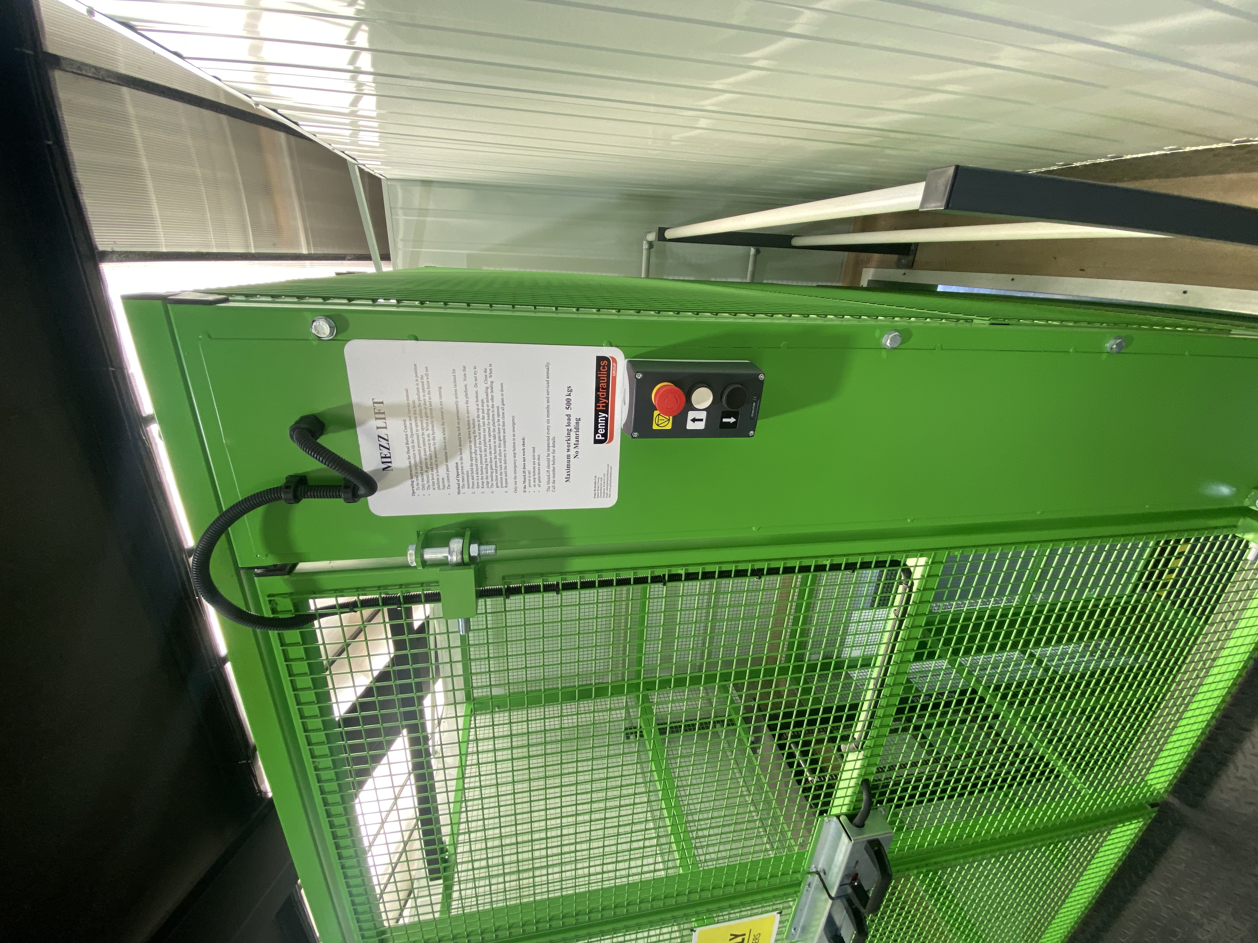 A green industrial mezzanine lift with a control panel, emergency stop button, and an instruction sheet attached to the cage; metal fencing surrounds the lift.
