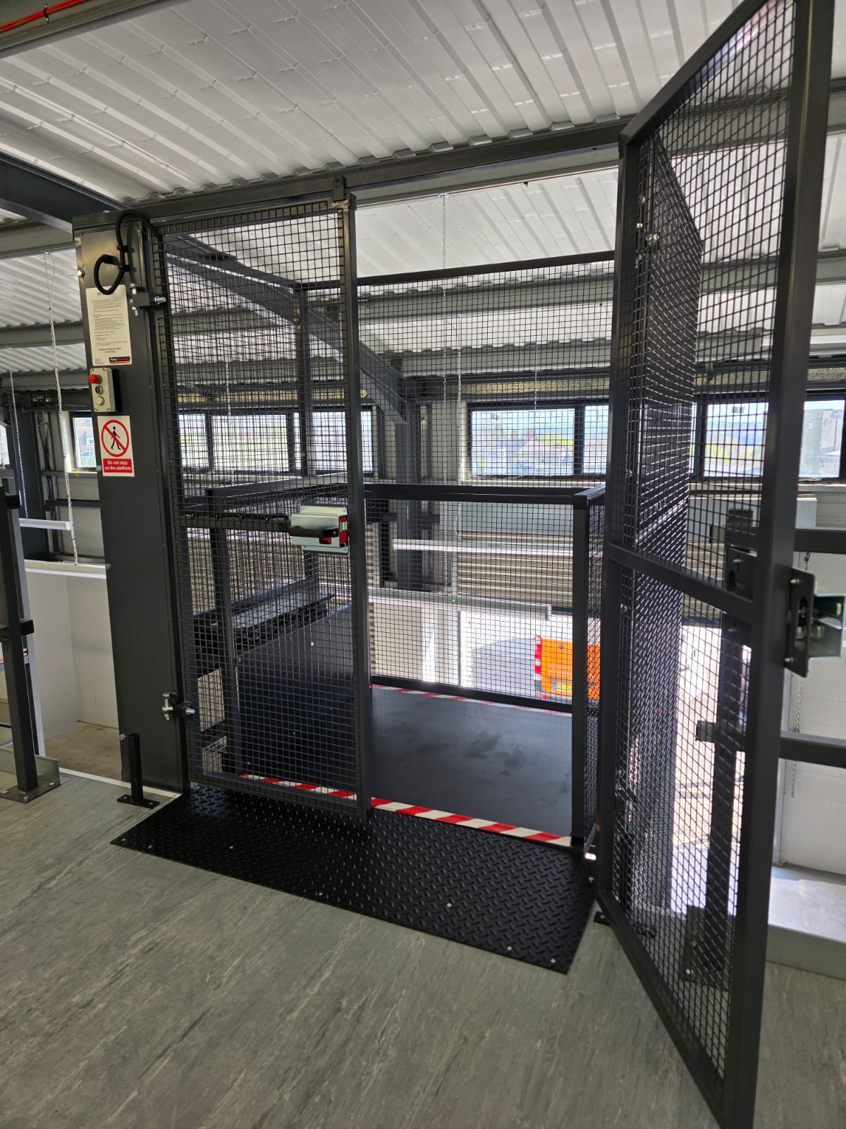 A large empty metal cage with multiple perches and platforms inside, viewed from outside through wire mesh. The structure appears to be an enclosure, possibly for animals.