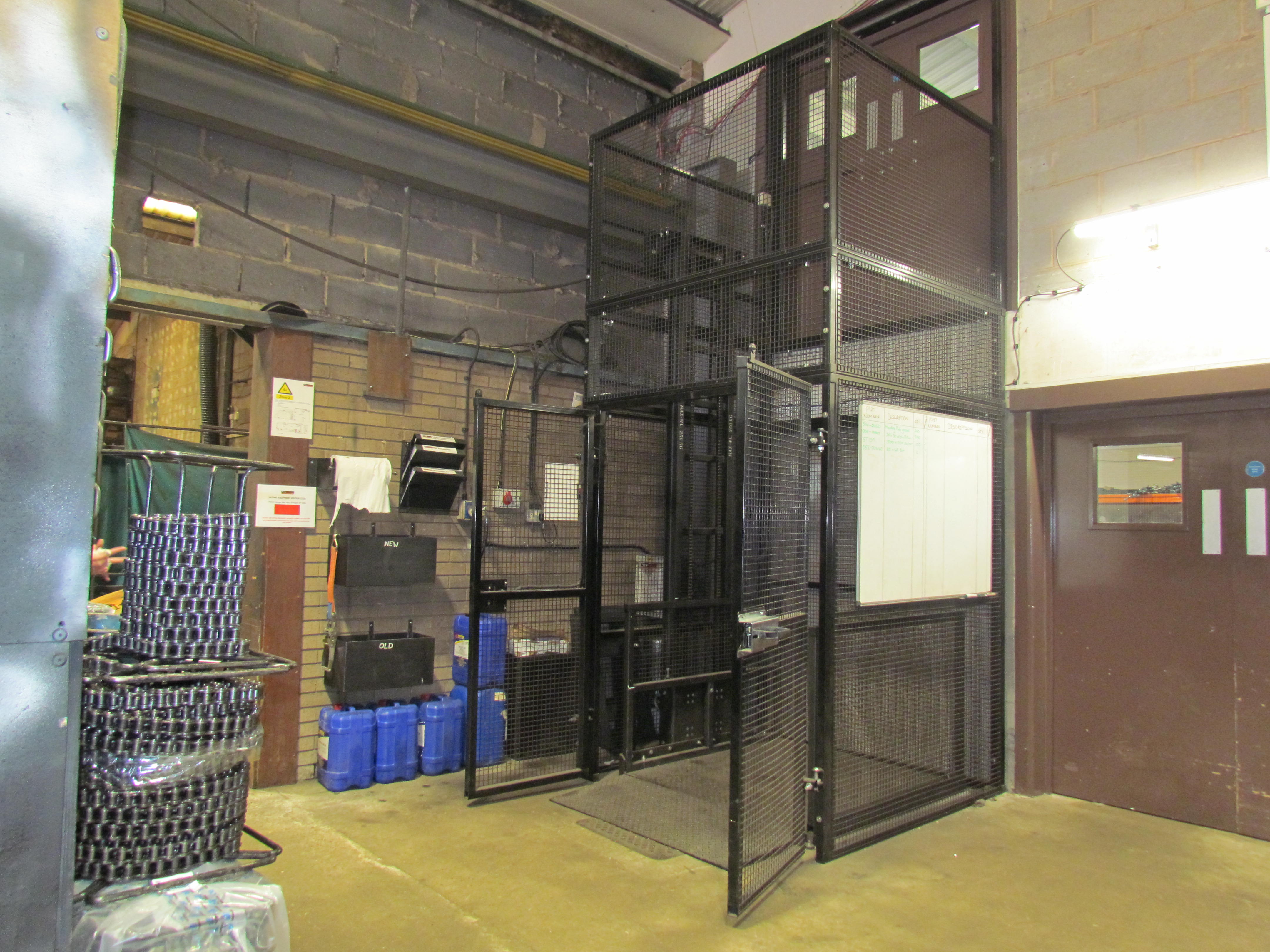 A metal goods lift with mesh fencing is installed in an industrial warehouse. Blue containers and storage bins are nearby, and a whiteboard is attached to the lift enclosure. The area has concrete floors and brick walls.