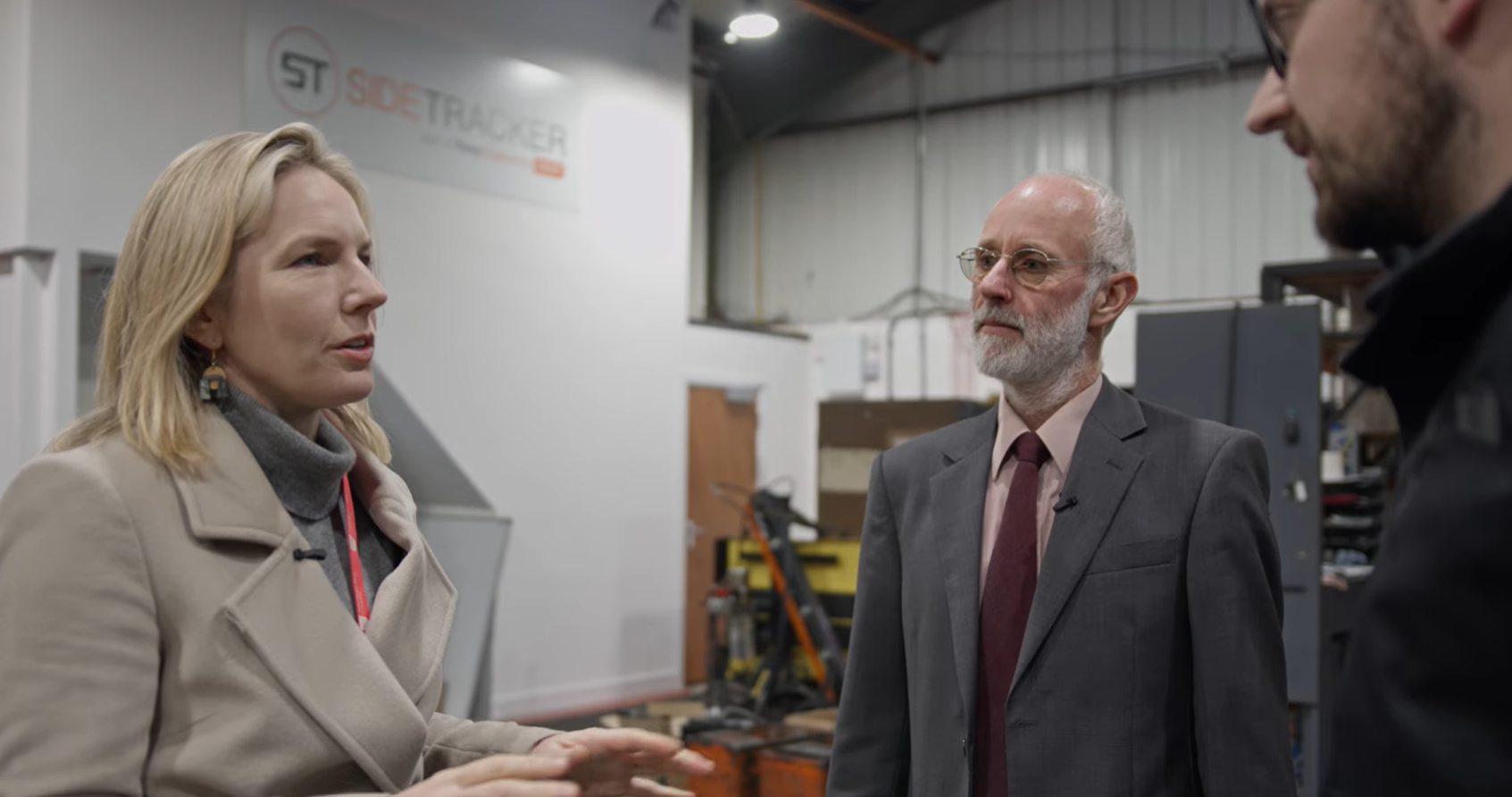 Three people stand talking in an industrial workspace. A woman gestures with her hands while speaking to two men, one of whom has a grey beard and wears a suit. Equipment and a SideTracker sign are visible in the background.