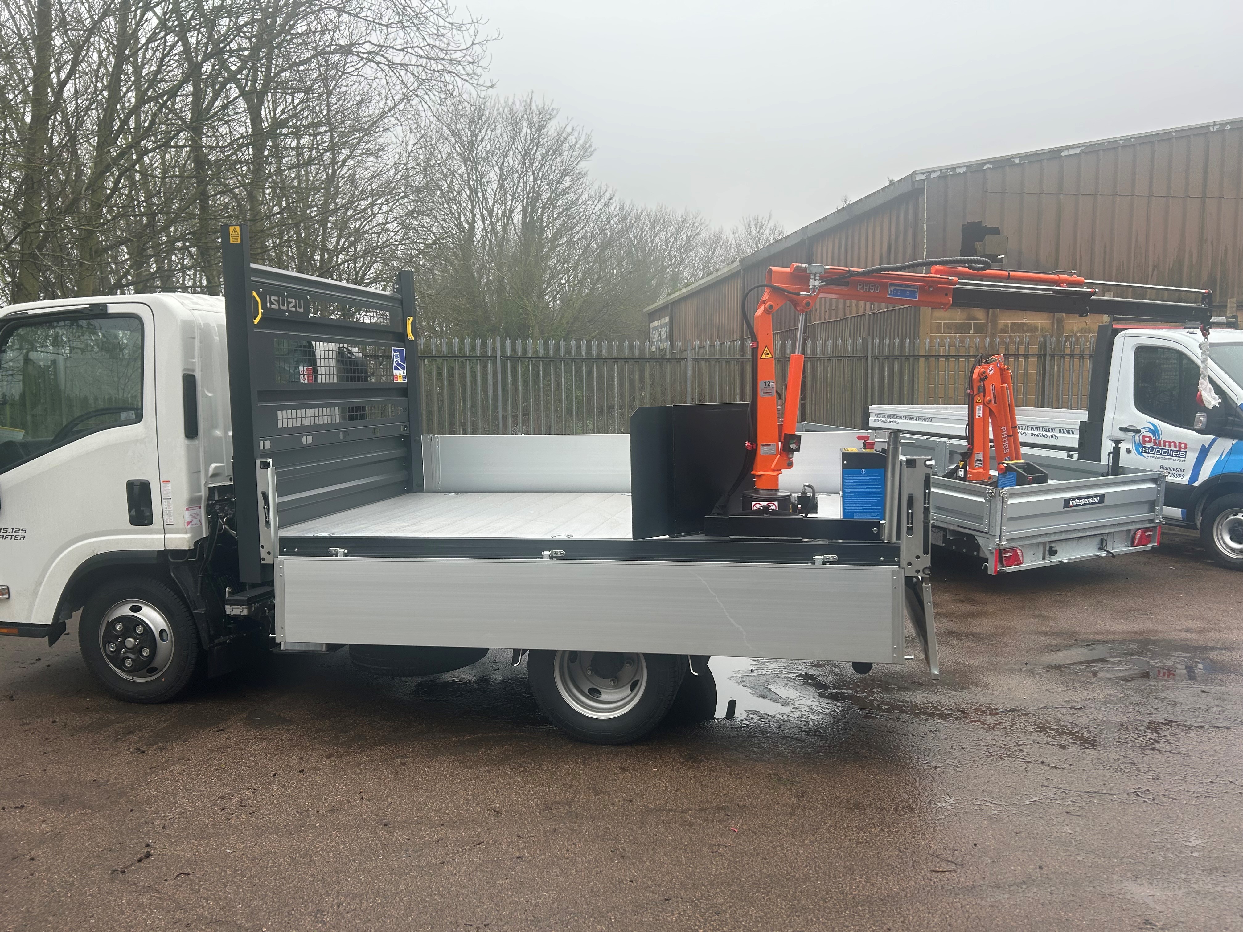A flatbed lorry with a mounted orange crane arm is parked outdoors on wet tarmac, next to another similar lorry; trees and a metal fence are visible in the background on a cloudy day.