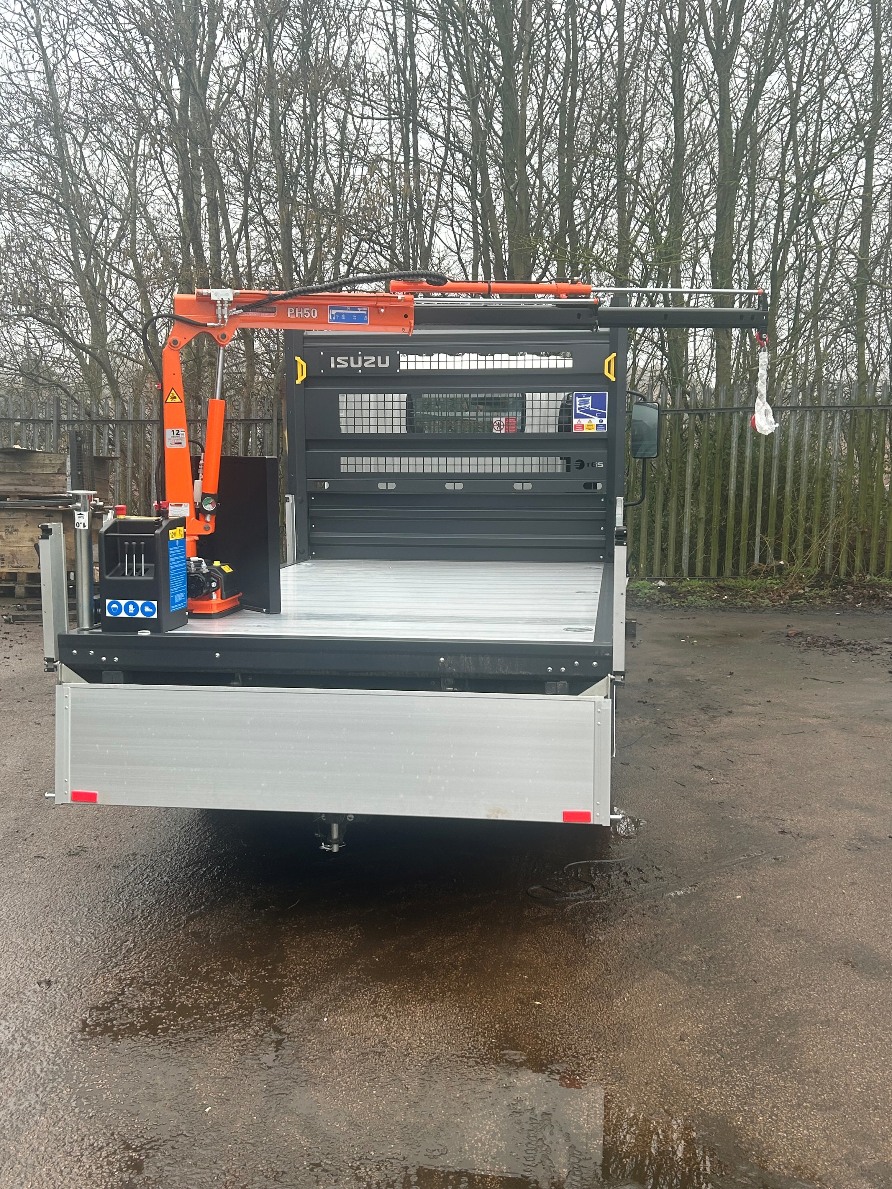 A flatbed lorry with an orange hydraulic crane mounted on the left side is parked on wet ground, with trees and a metal fence in the background.