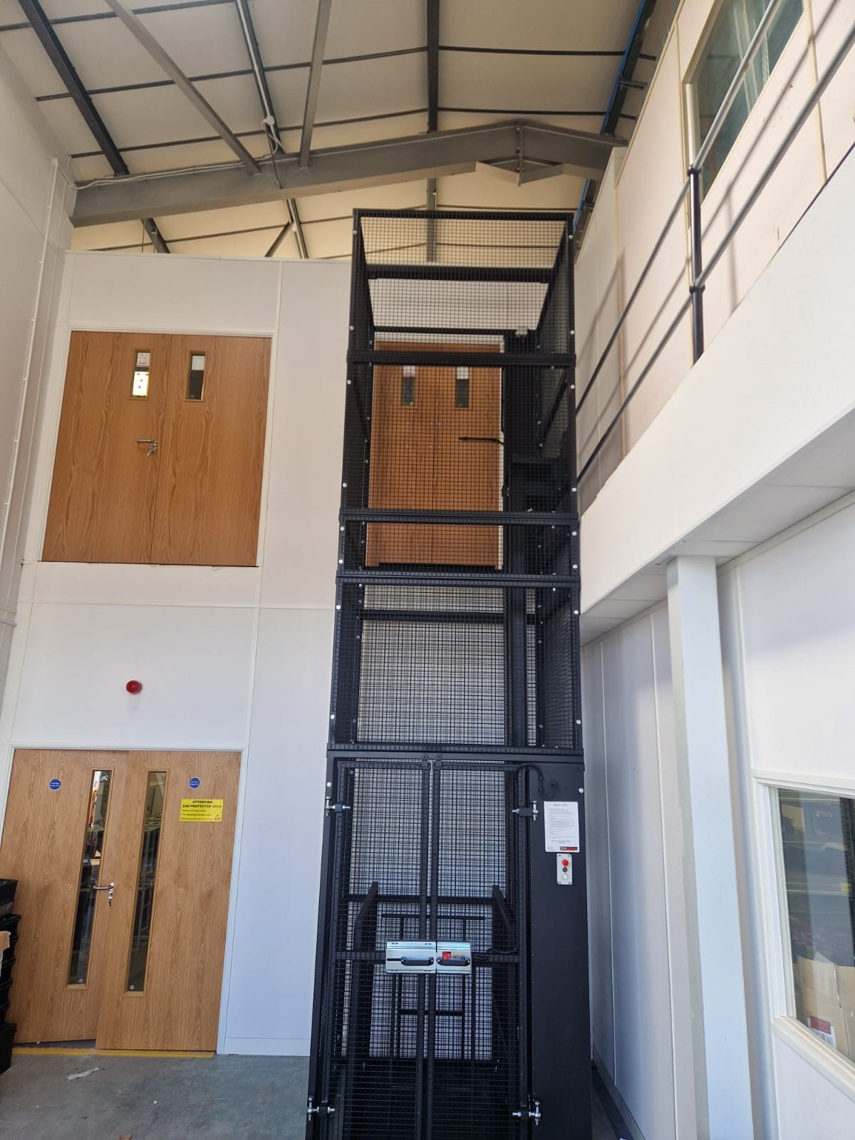 A two-storey industrial interior with wooden doors on both levels, featuring a black metal goods lift cage in the centre, and white walls with windows and a steel-railed walkway on the upper floor.
