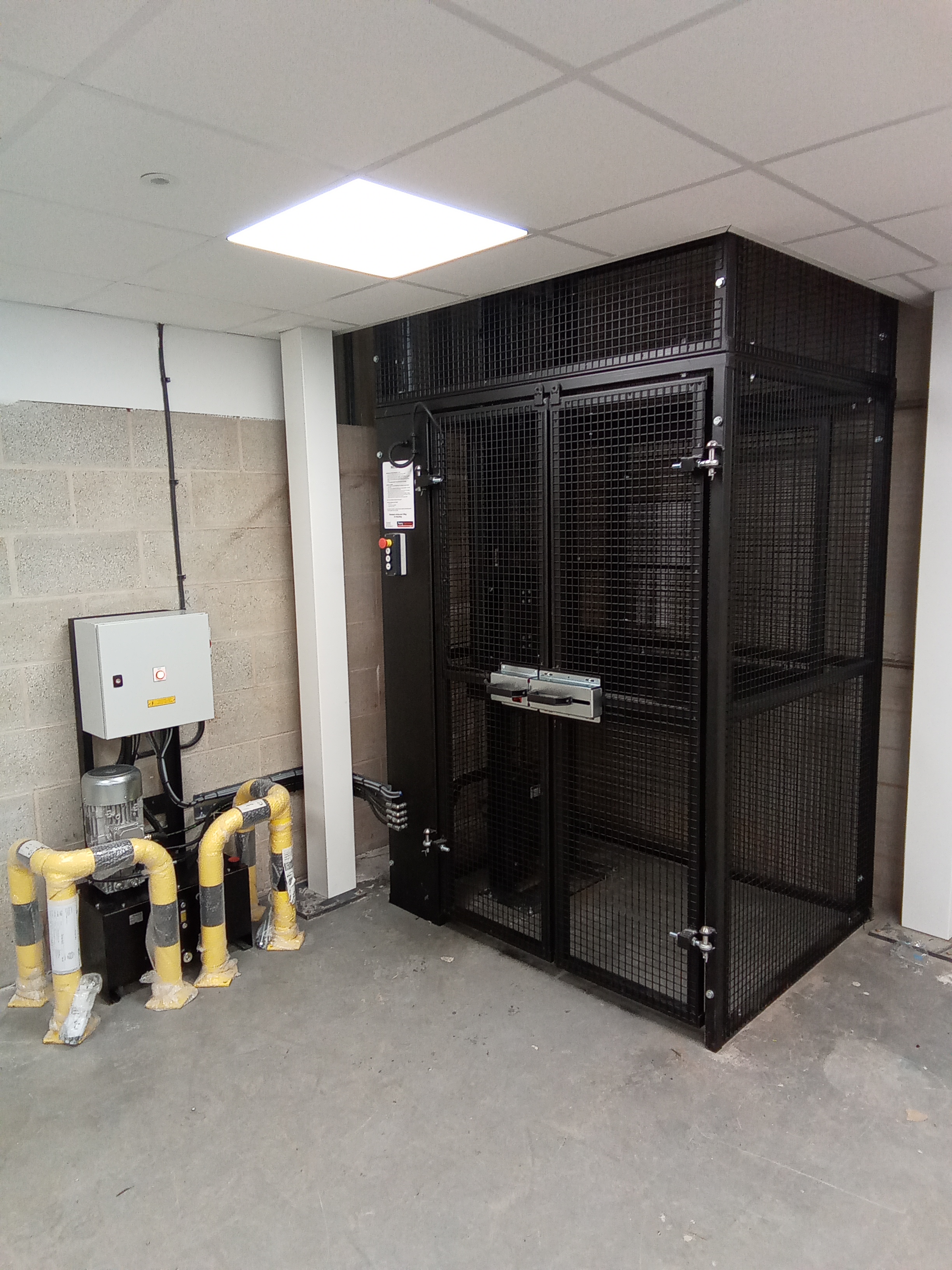 A secure, black metal cage enclosure is installed in the corner of a room with a concrete floor and breeze block walls. Nearby are exposed pipes wrapped in yellow insulation and an electrical control panel.