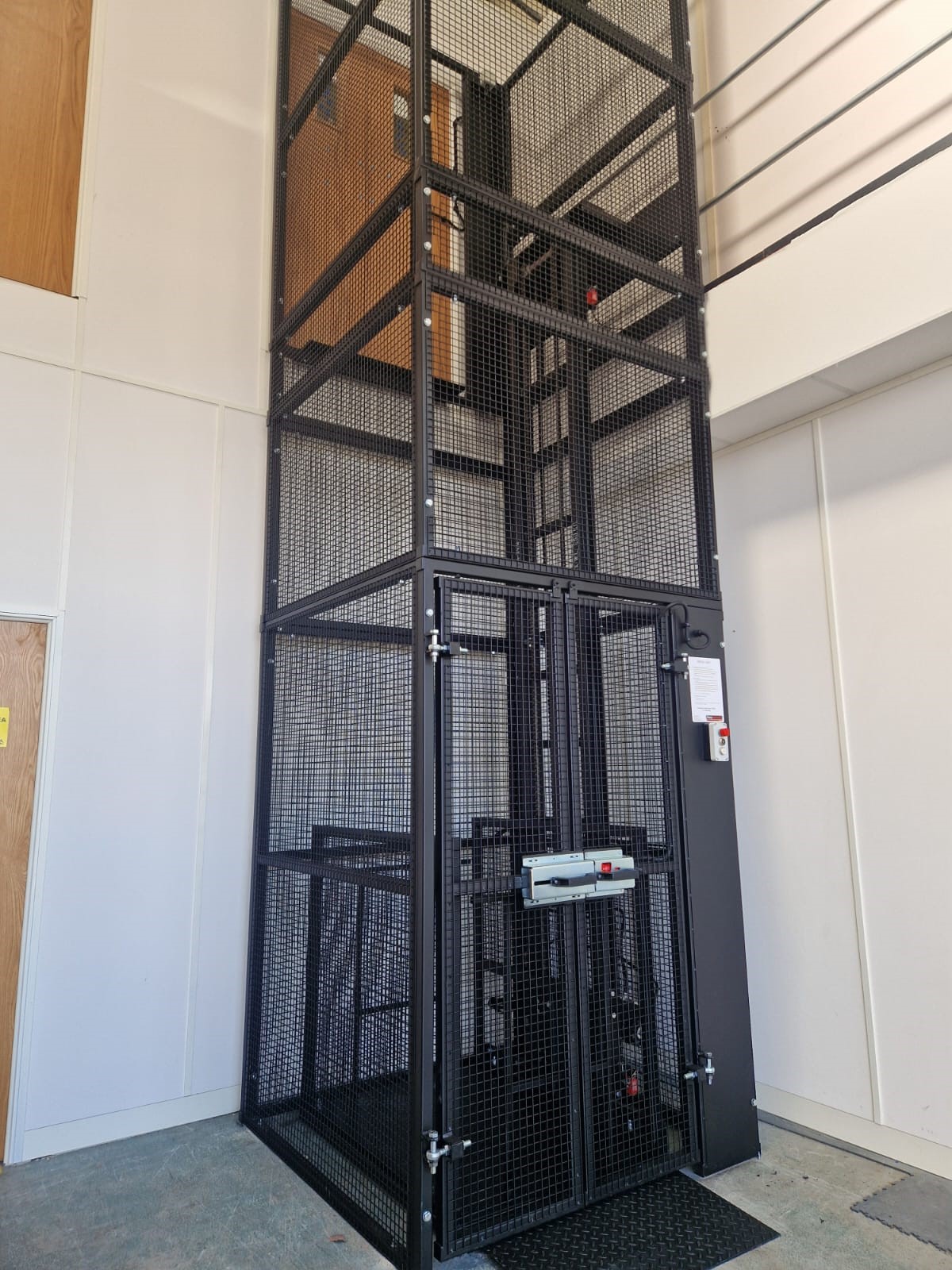 A black metal cage lift with mesh sides is installed inside a white room, reaching up to the upper floor and a wooden door at the top. The lift has control buttons on the front.
