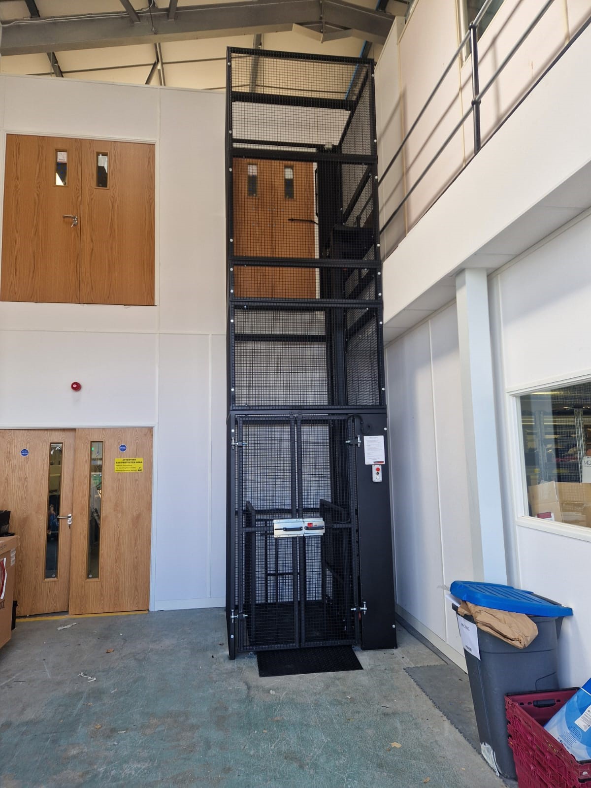 A black metal goods lift with mesh sides stands in a warehouse-style room with white walls, two sets of wooden doors, and a blue recycling bin in the corner. The lift reaches up to a first-floor balcony.
